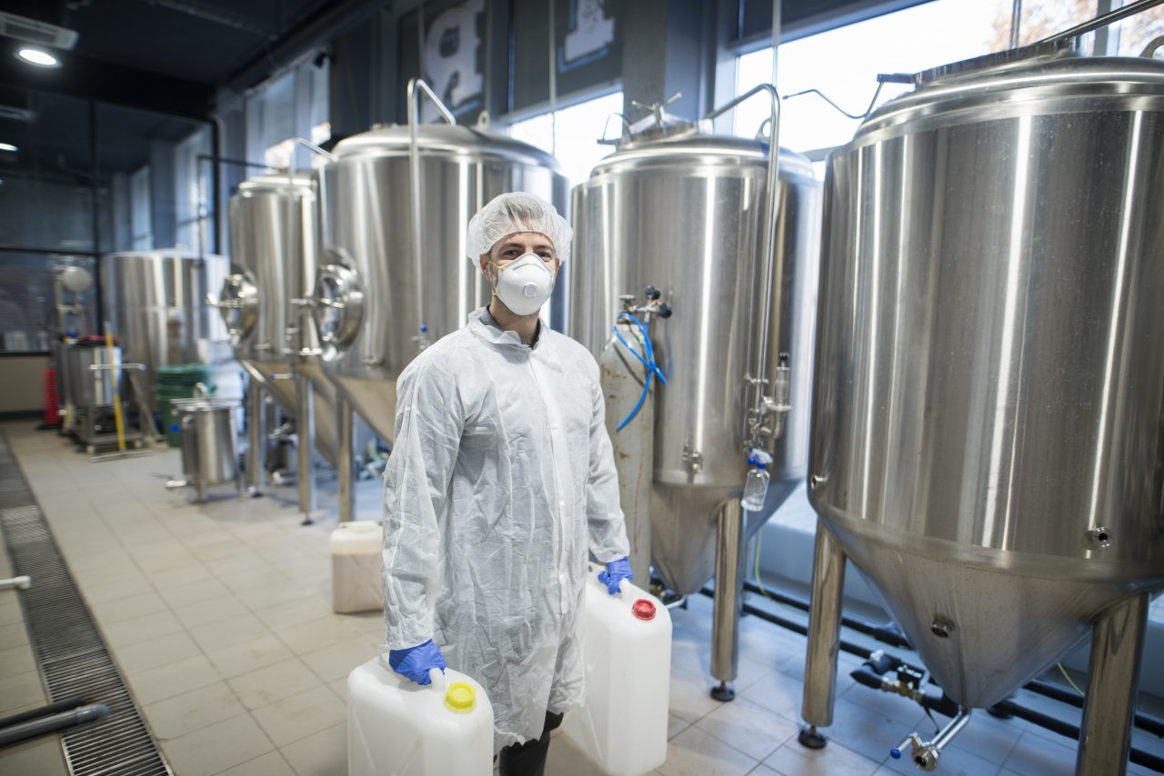 Industrial Worker Technologist In White Protective Suit With Hairnet And Mask Holding Plastic Cans With Chemicals In Food Factory Production Line.